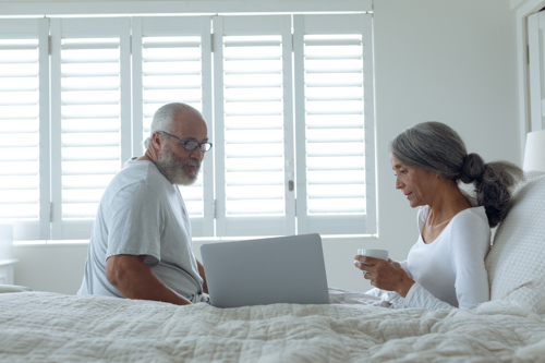 Side view of senior diverse couple sitting on bed inside a room.